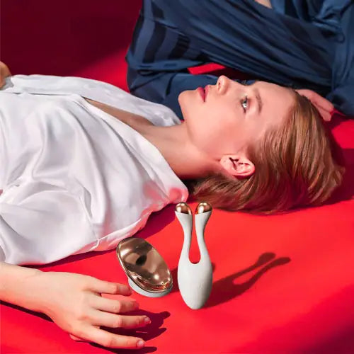 "White RF facial device with gold accents, placed beside a woman lying on a red surface, wearing a white blouse."
