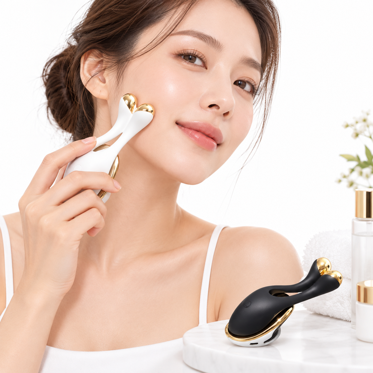 A woman with a clean complexion uses a white and gold facial roller on her cheek. Another black and gold facial roller rests on a marble tray beside her, with skincare bottles and a small plant in the background against a white studio backdrop.