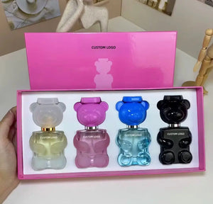"Four mini bear-shaped perfume bottles in pink, blue, clear, and black, displayed in a pink gift box."