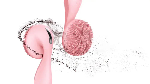 Pink sonic facial cleansing brush with textured silicone bristles, featuring a curved handle and water droplets around it.