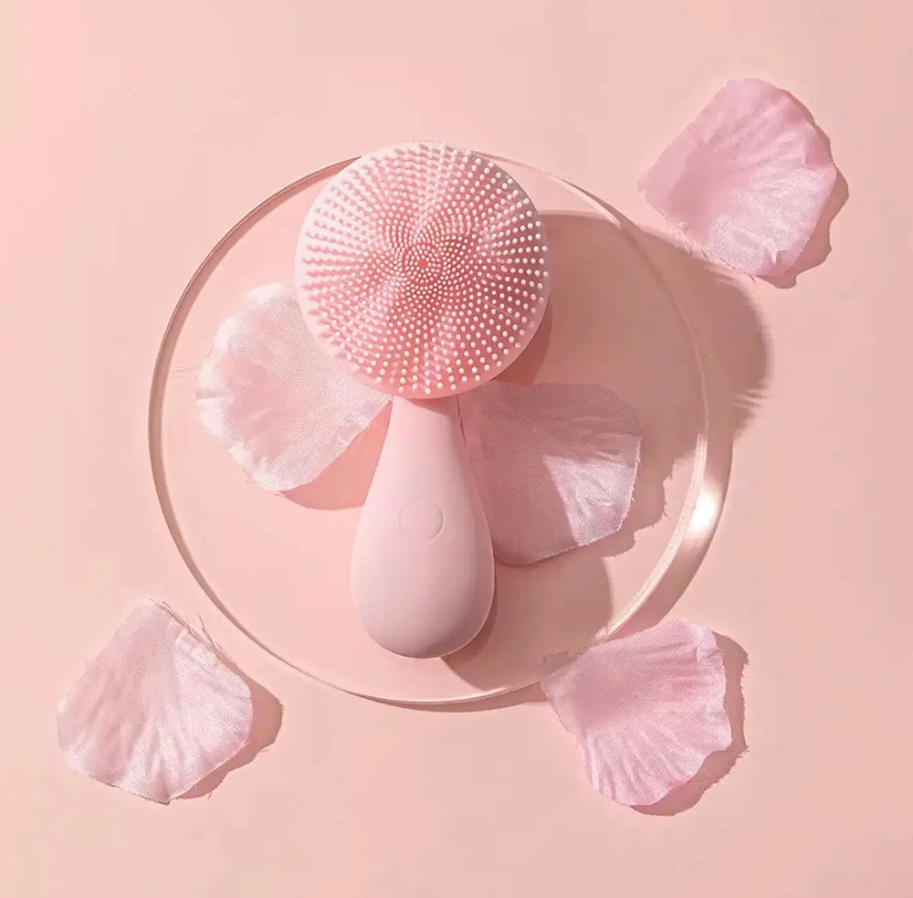 Pink sonic facial cleansing brush with soft silicone bristles, placed on a clear circular dish, surrounded by pink rose petal