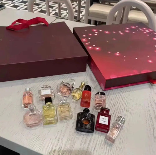 "Maroon gift box with red ribbon, open to reveal 12 mini perfume bottles in various shapes and colors on a white wooden table