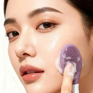 "Purple double-ended silicone exfoliating brush held against cheek, with foam cleanser visible on the brush surface."